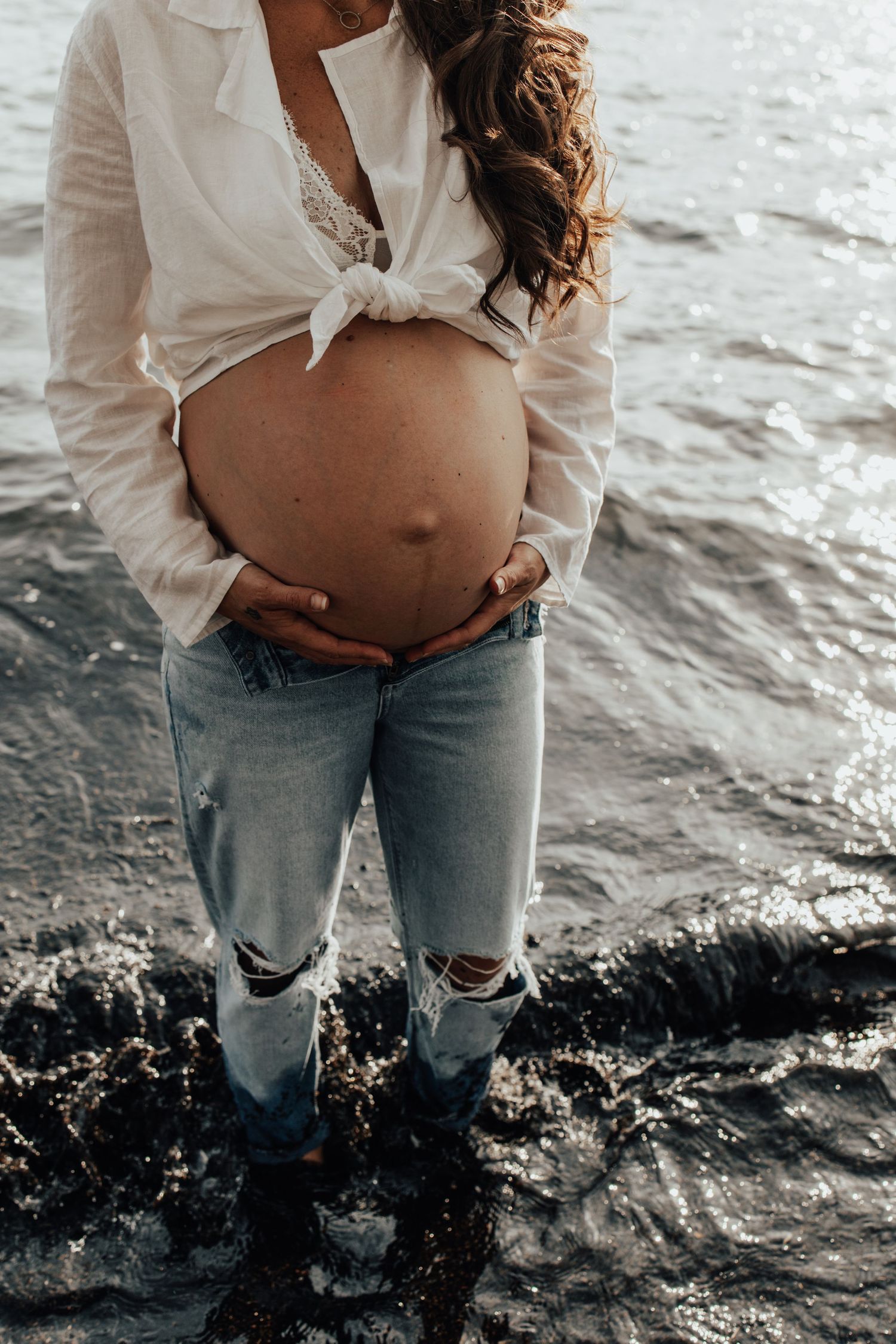 Maternity photo showing pregnant belly in white tied shirt and distressed jeans by the water.