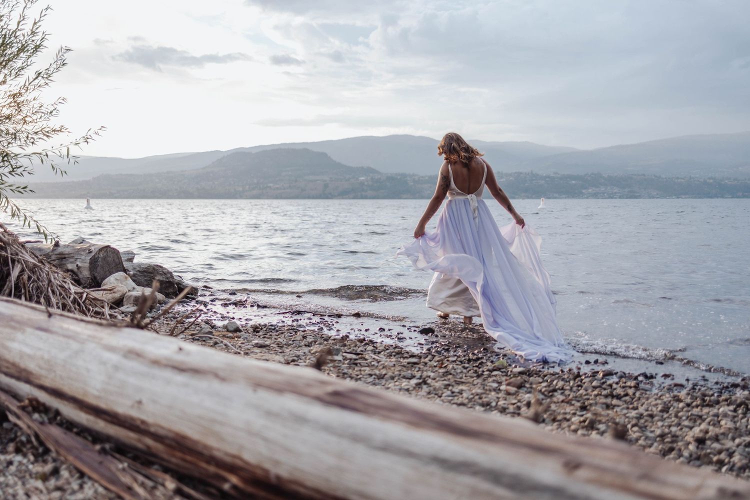 Person in white dress exploring rocky shoreline with mountains in background.