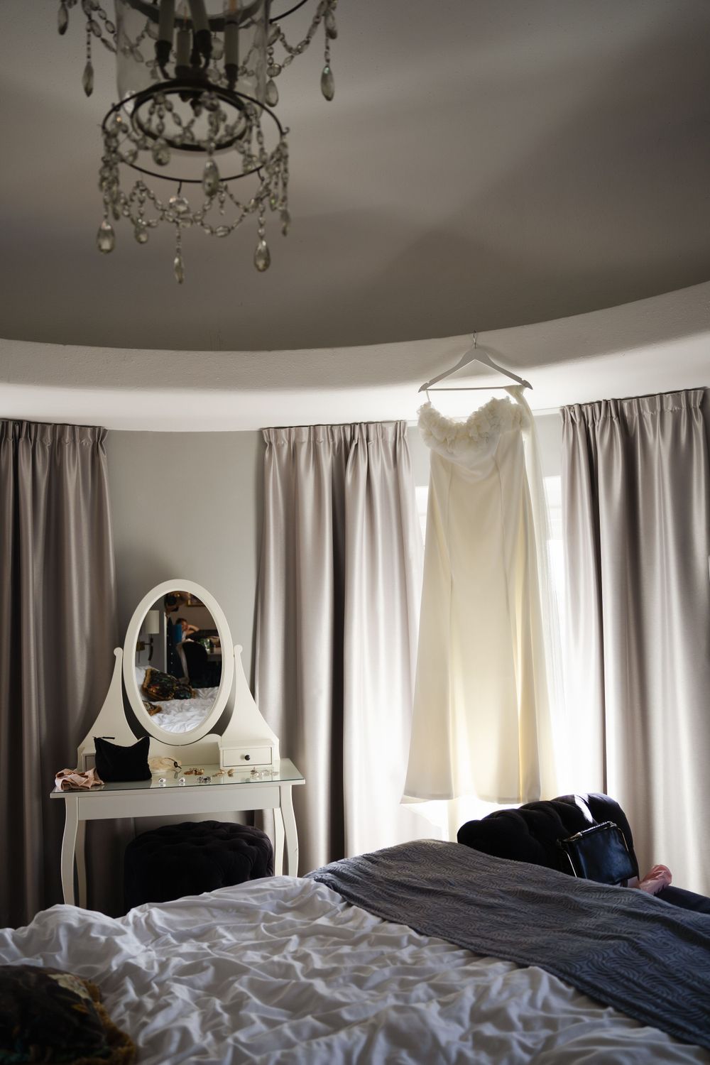 A wedding dress hangs in an elegant bedroom with crystal chandelier and vanity mirror.