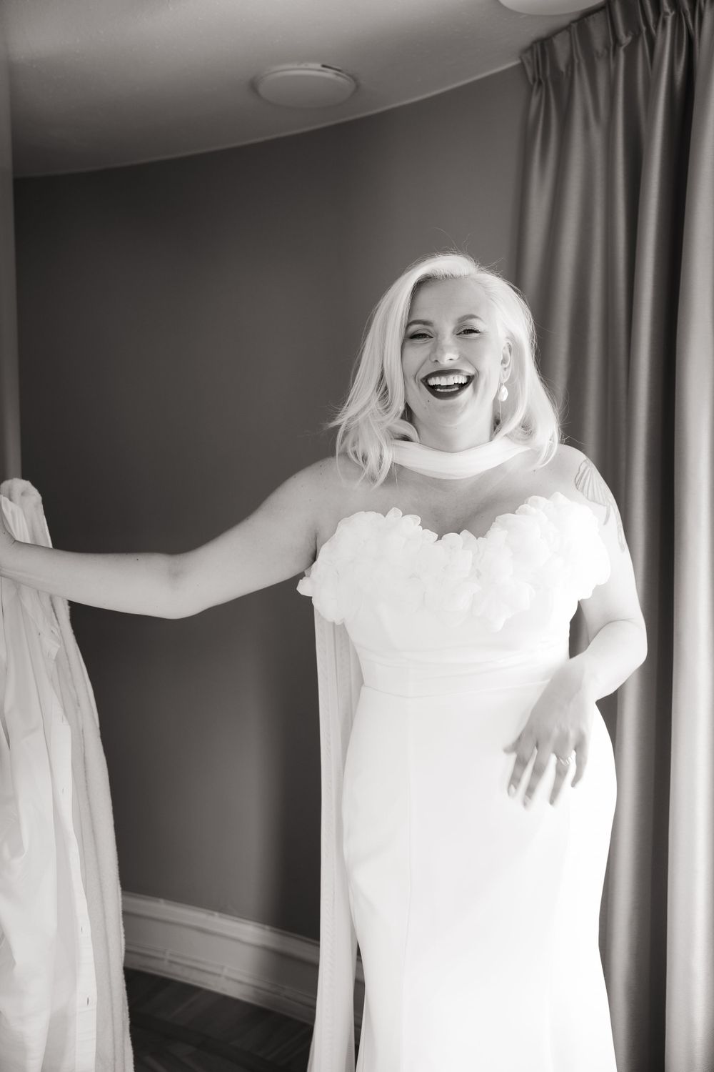 Bride in white strapless wedding dress smiling and posing against gray wall with curtains.
