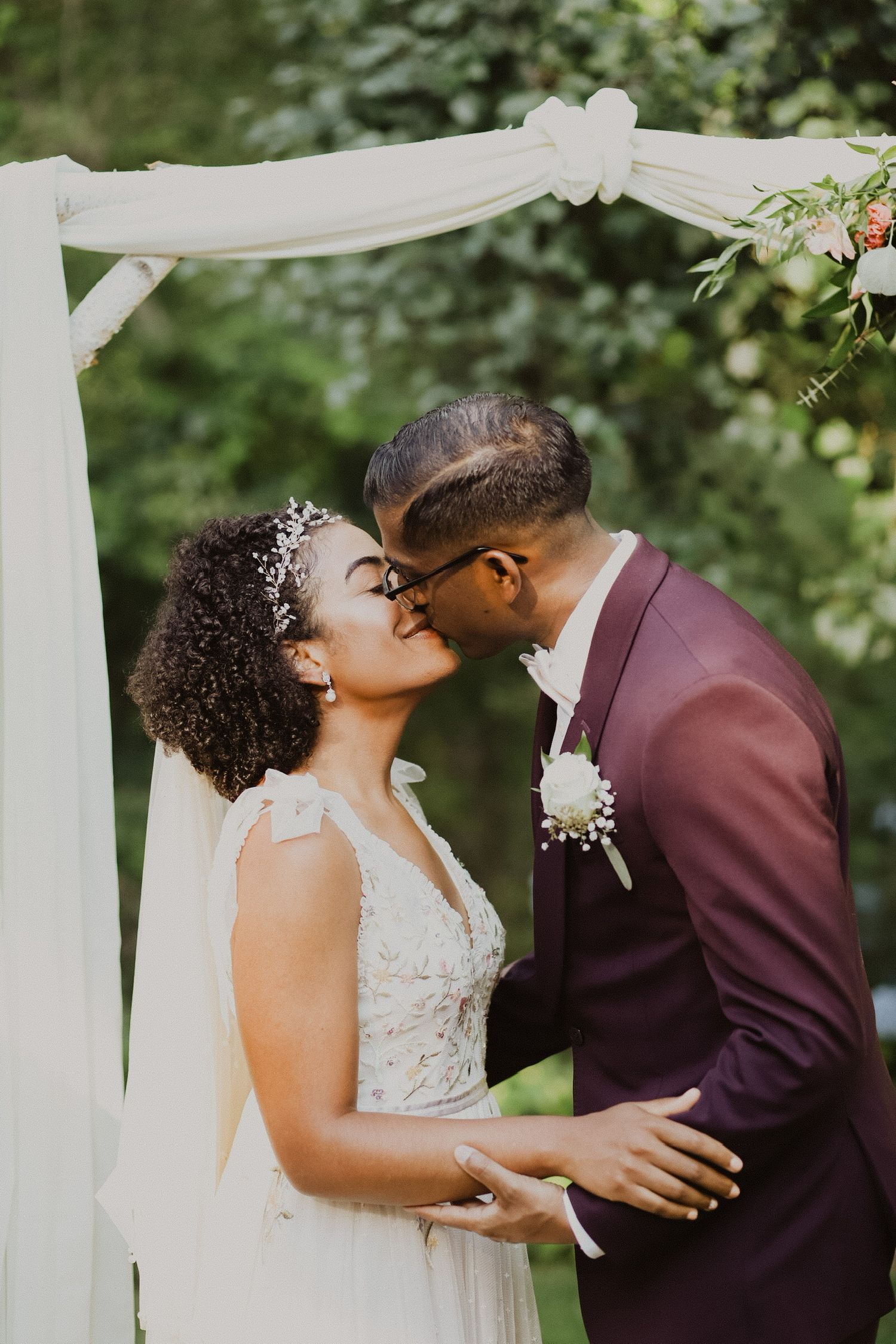 Newlyweds share their first kiss under white draped wedding arch in garden setting.