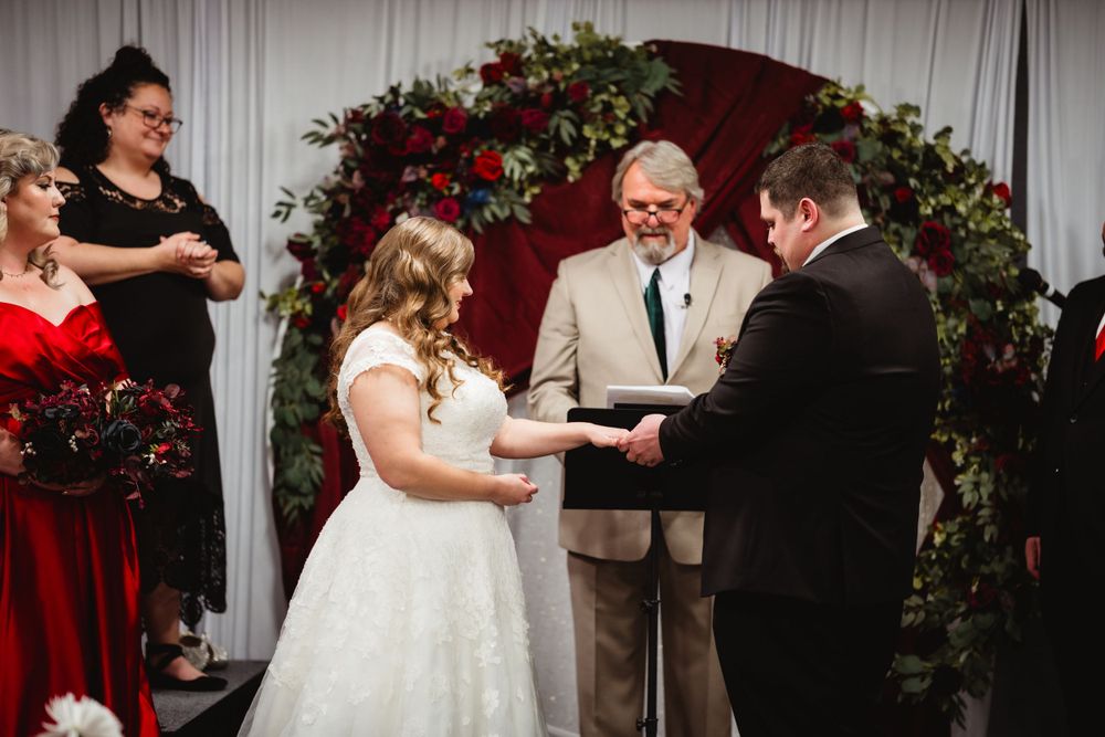 Wedding ceremony in progress under floral arch with red and white decor.