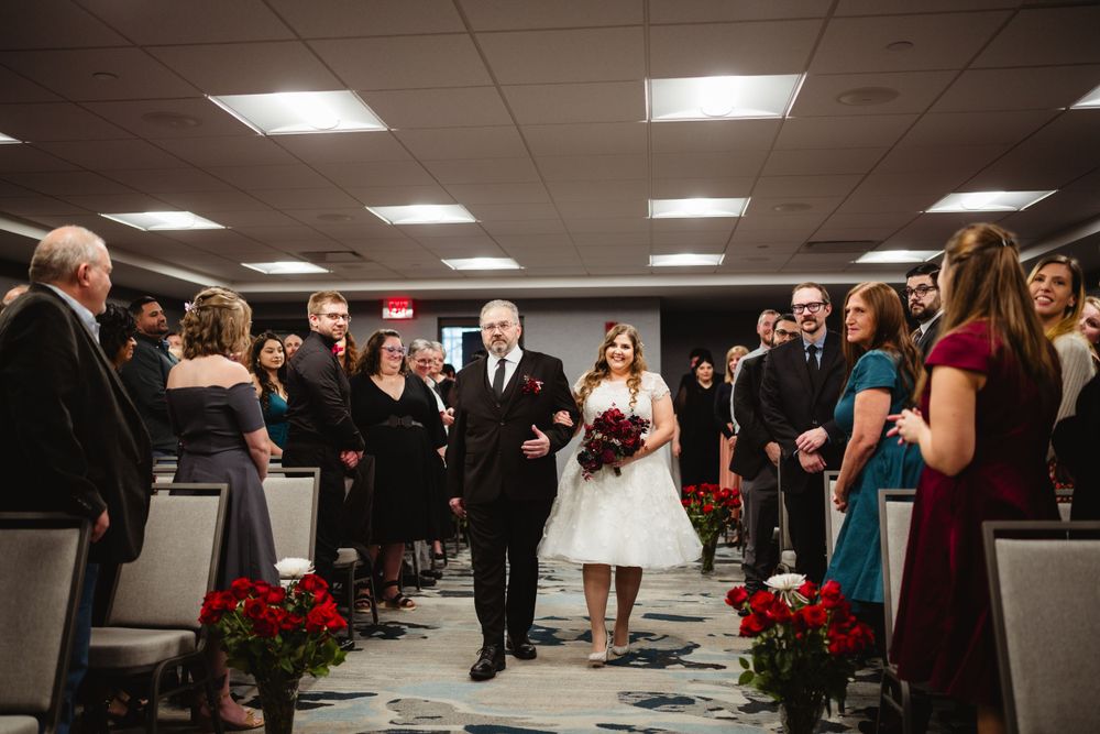 Wedding processional in indoor venue with red floral decorations and lined chairs.