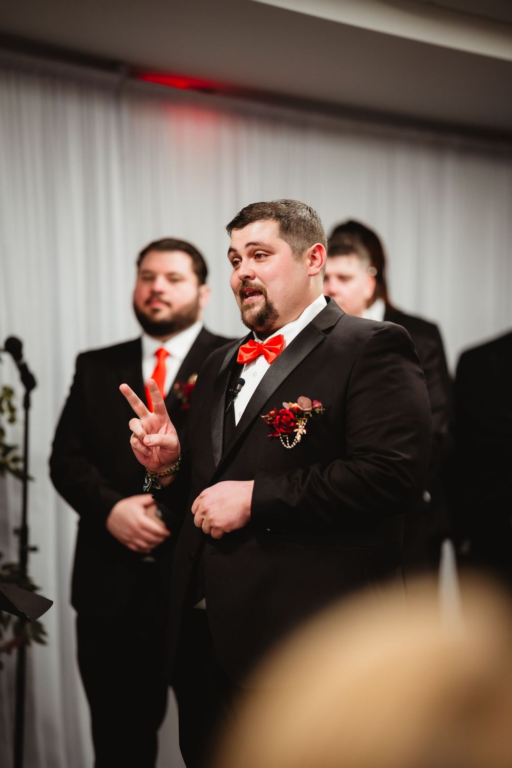 Member of wedding party in black suit and red bowtie gestures during indoor ceremony.