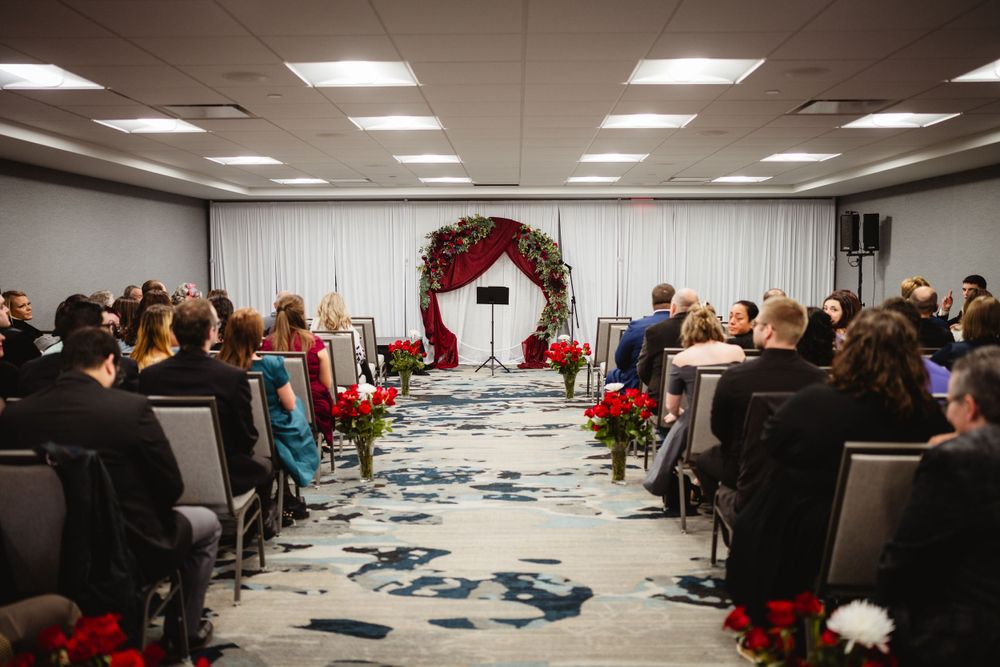 Wedding ceremony venue setup with red floral arch and rows of chairs on patterned carpet.