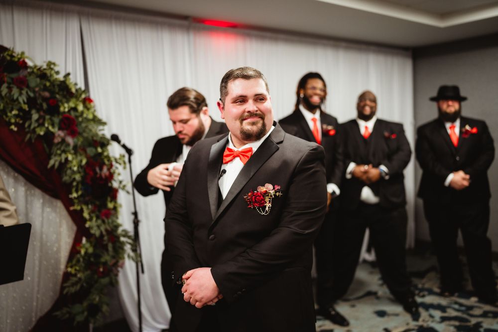 Wedding party member in black suit and red bowtie stands in ceremony venue.