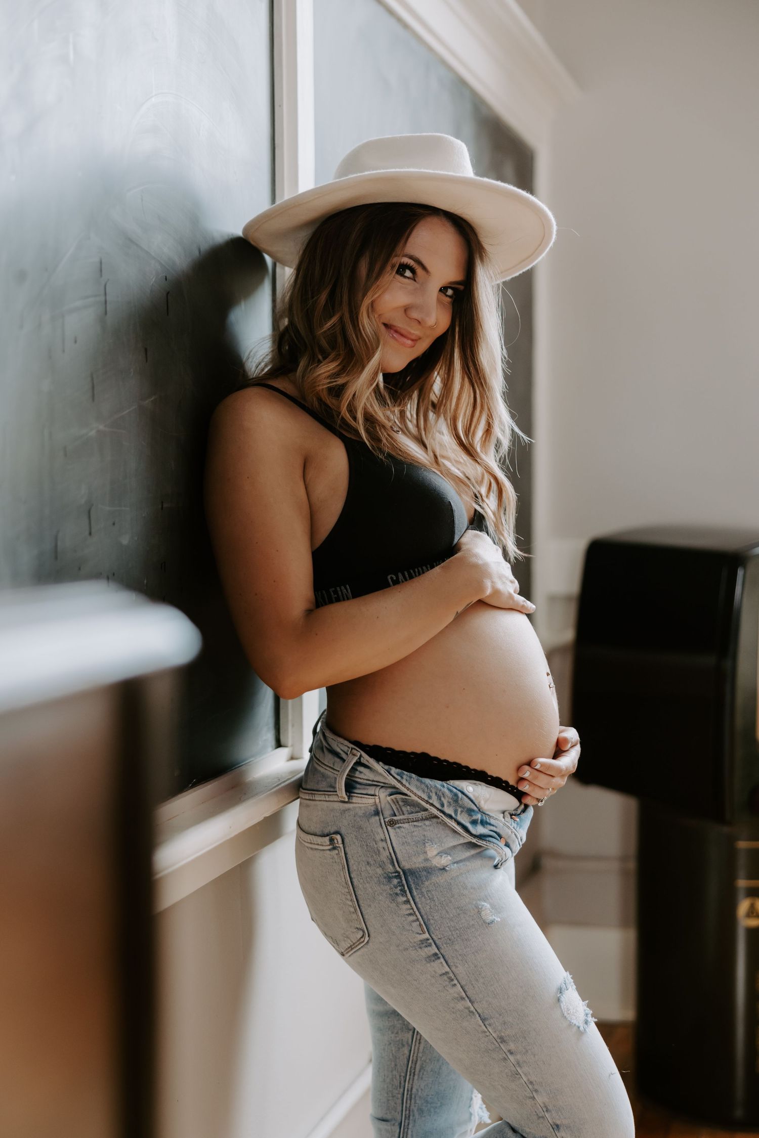 Stylish pregnant woman in black crop top, jeans, and wide-brim hat cradles her baby bump indoors.