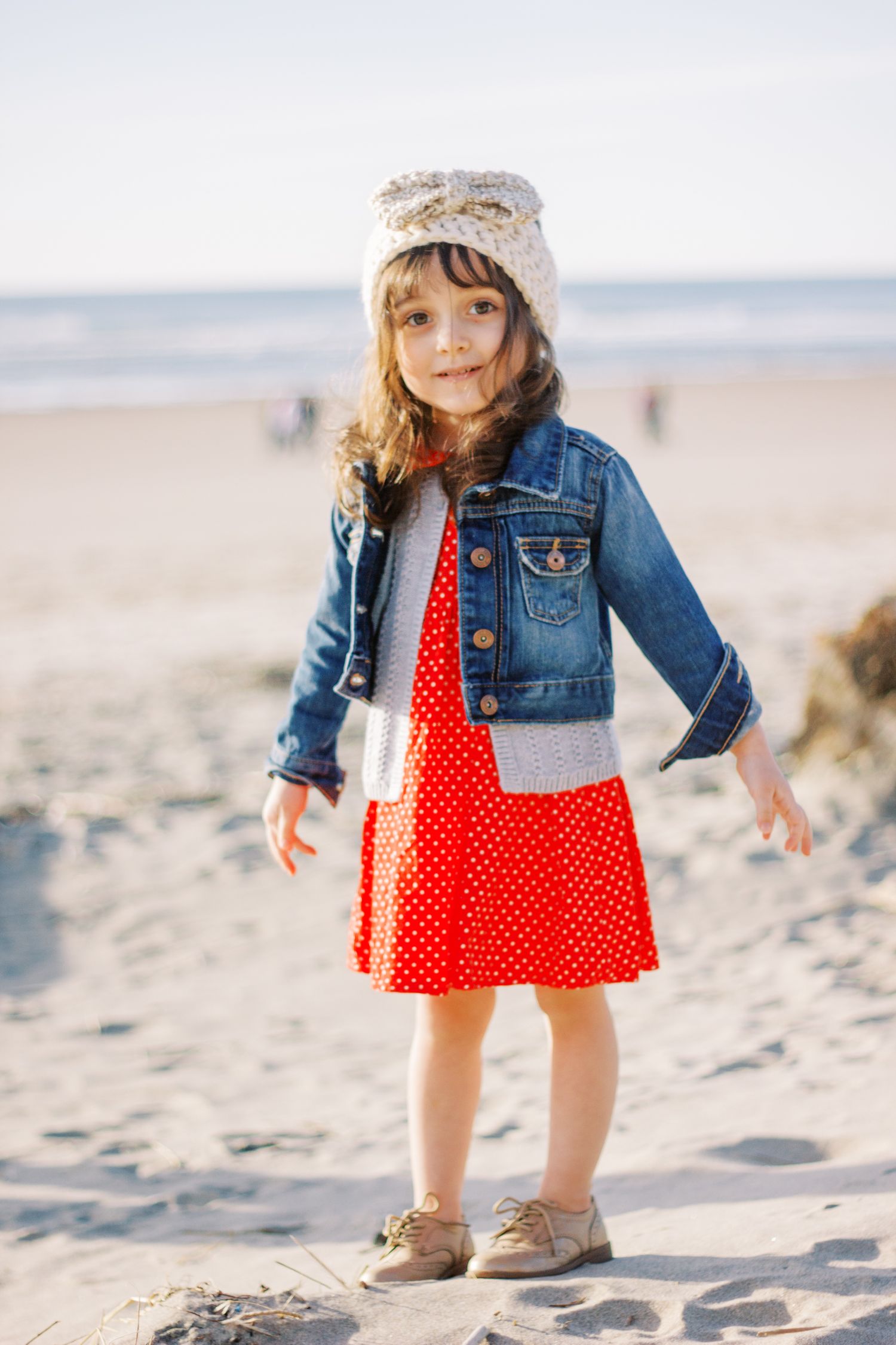 Child in denim jacket and red polka dot dress standing on sandy beach.
