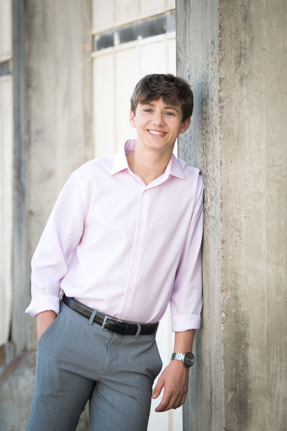 Portrait of a person in a pink dress shirt and gray slacks leaning against a concrete wall.