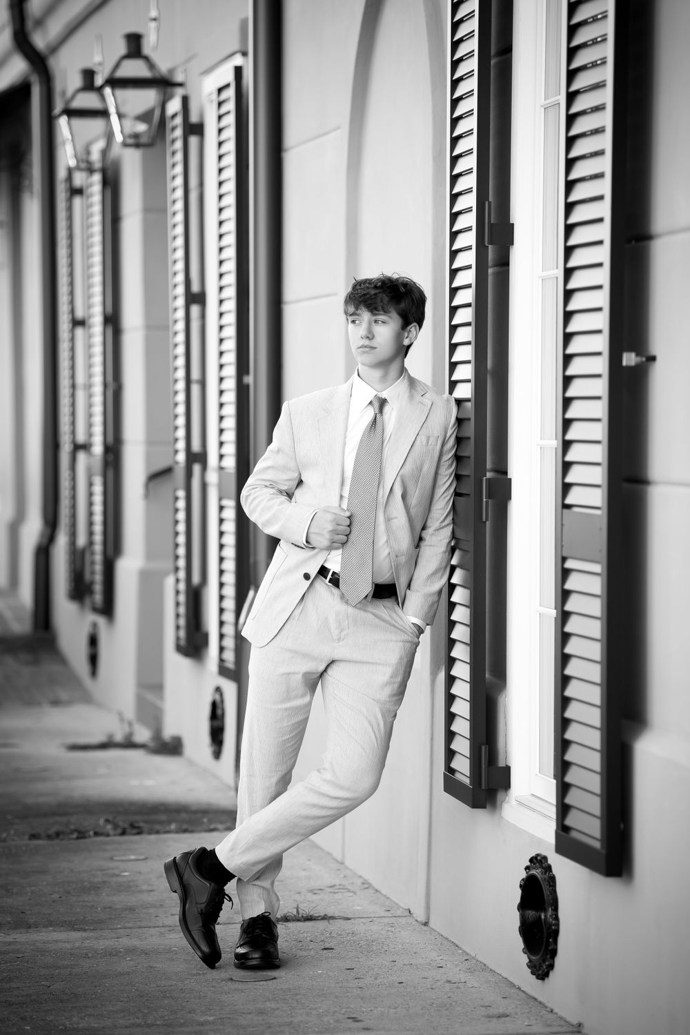 Black and white photo of a person in a light suit posing against shuttered windows with vintage street lamps.