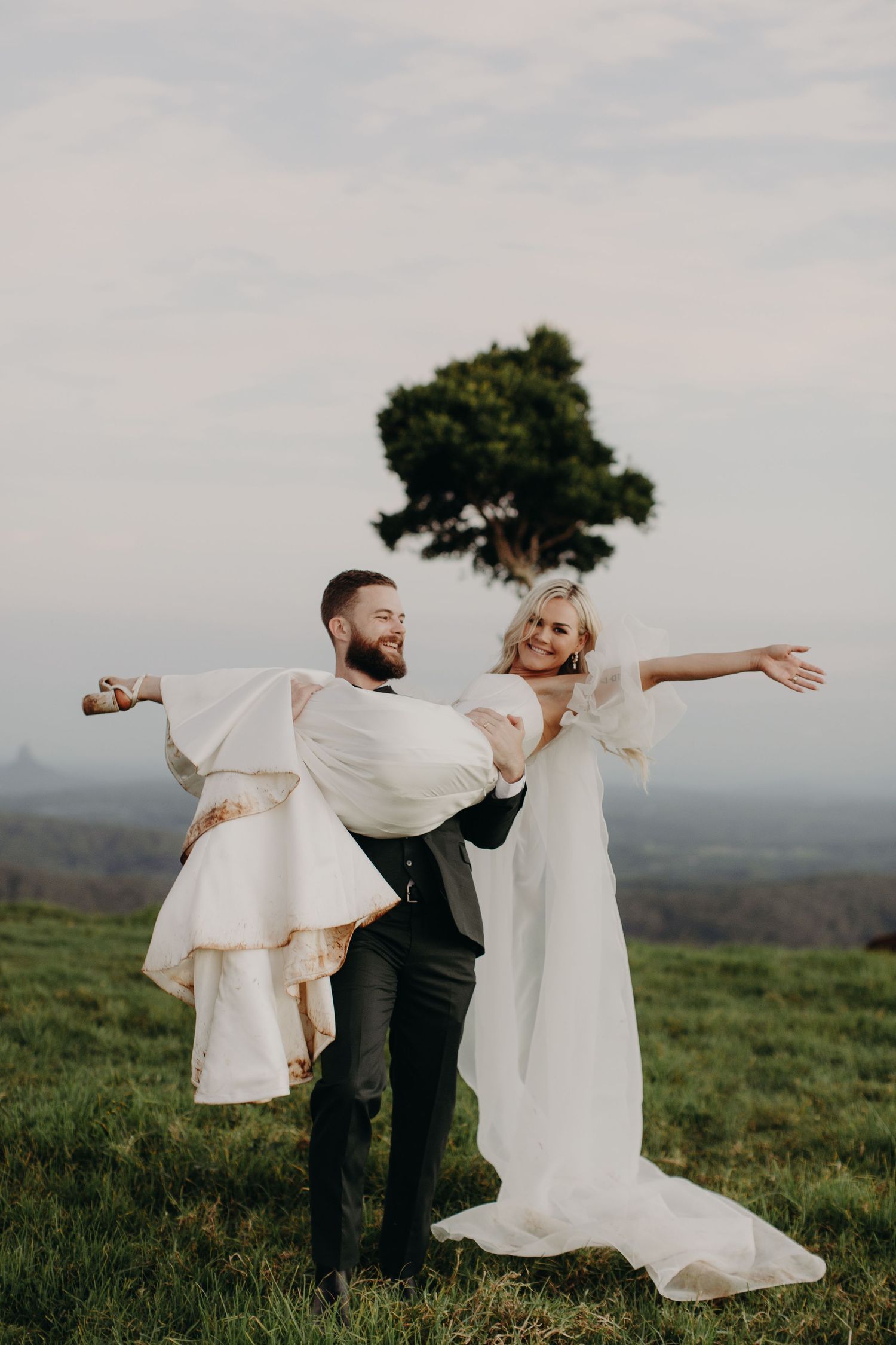 Newlyweds playfully posing with outstretched arms in a grassy hilltop field.