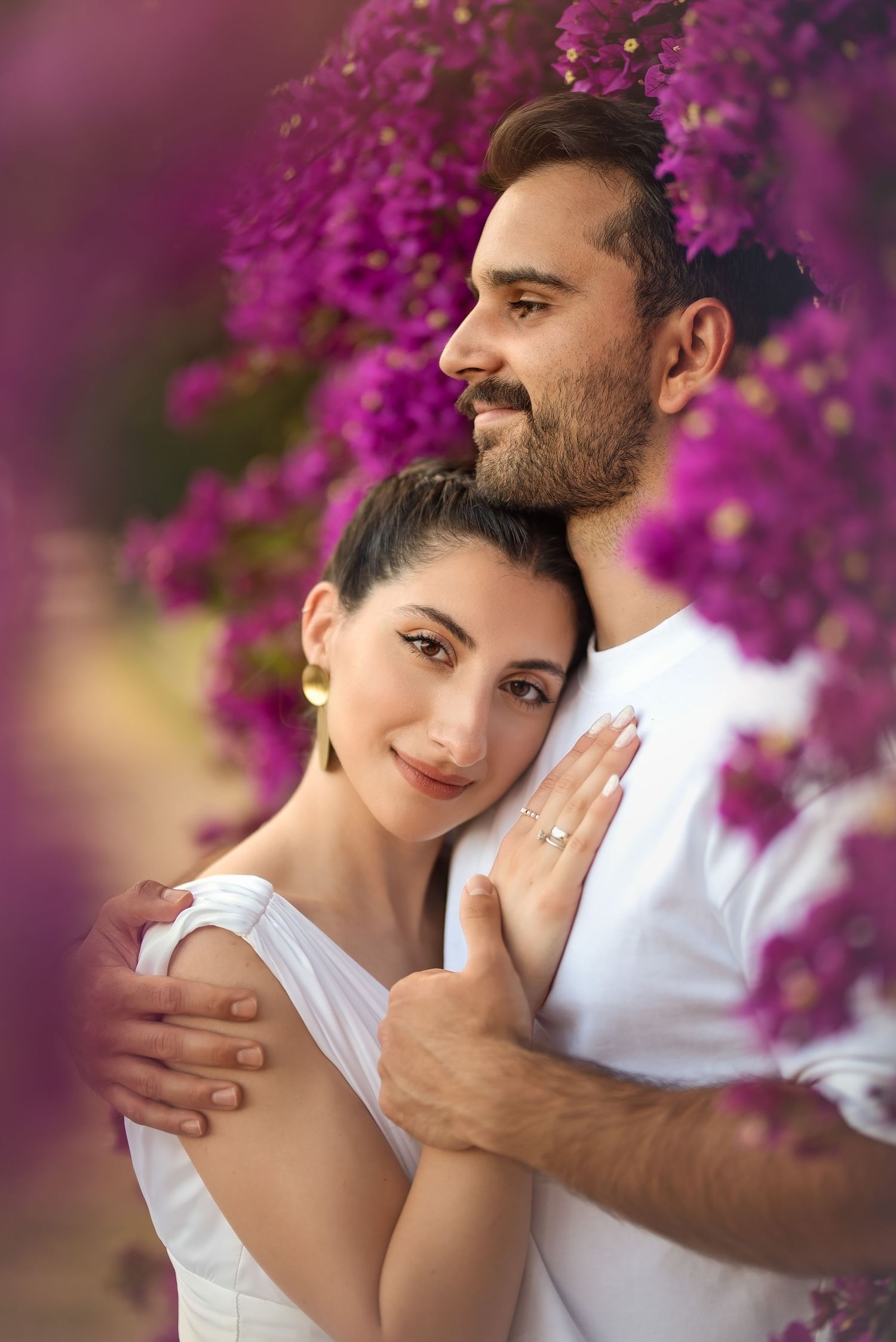 Romantic couple embraces among vibrant purple bougainvillea flowers in soft lighting.