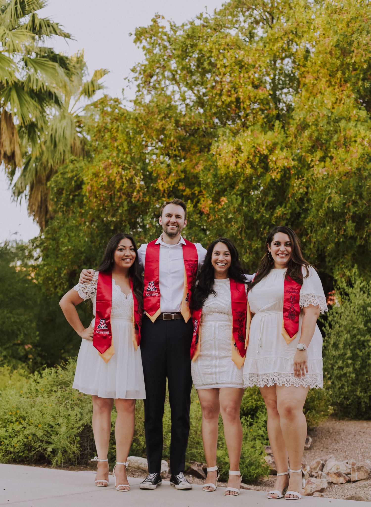 A graduation group photo showing four people in white outfits with red stoles standing under trees.