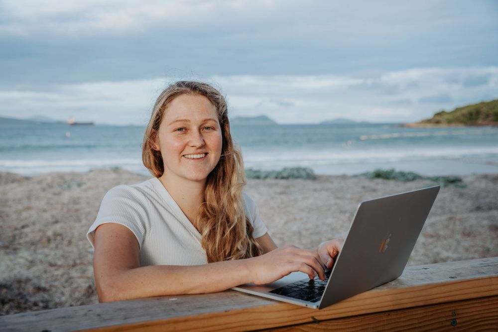 Person lying on beach using laptop with ocean waves in background.