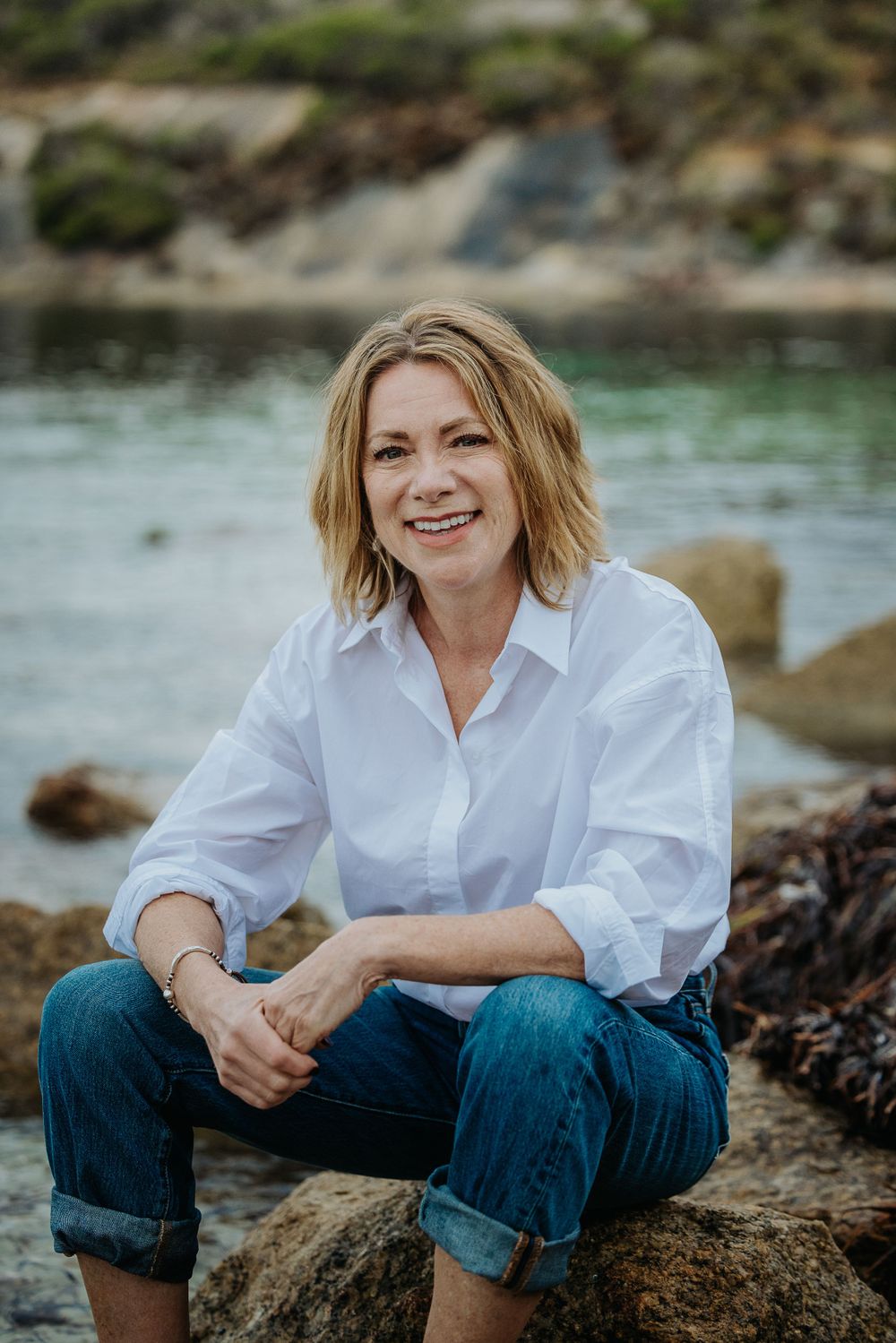 Person in white shirt and jeans sitting casually on coastal rocks by the water.