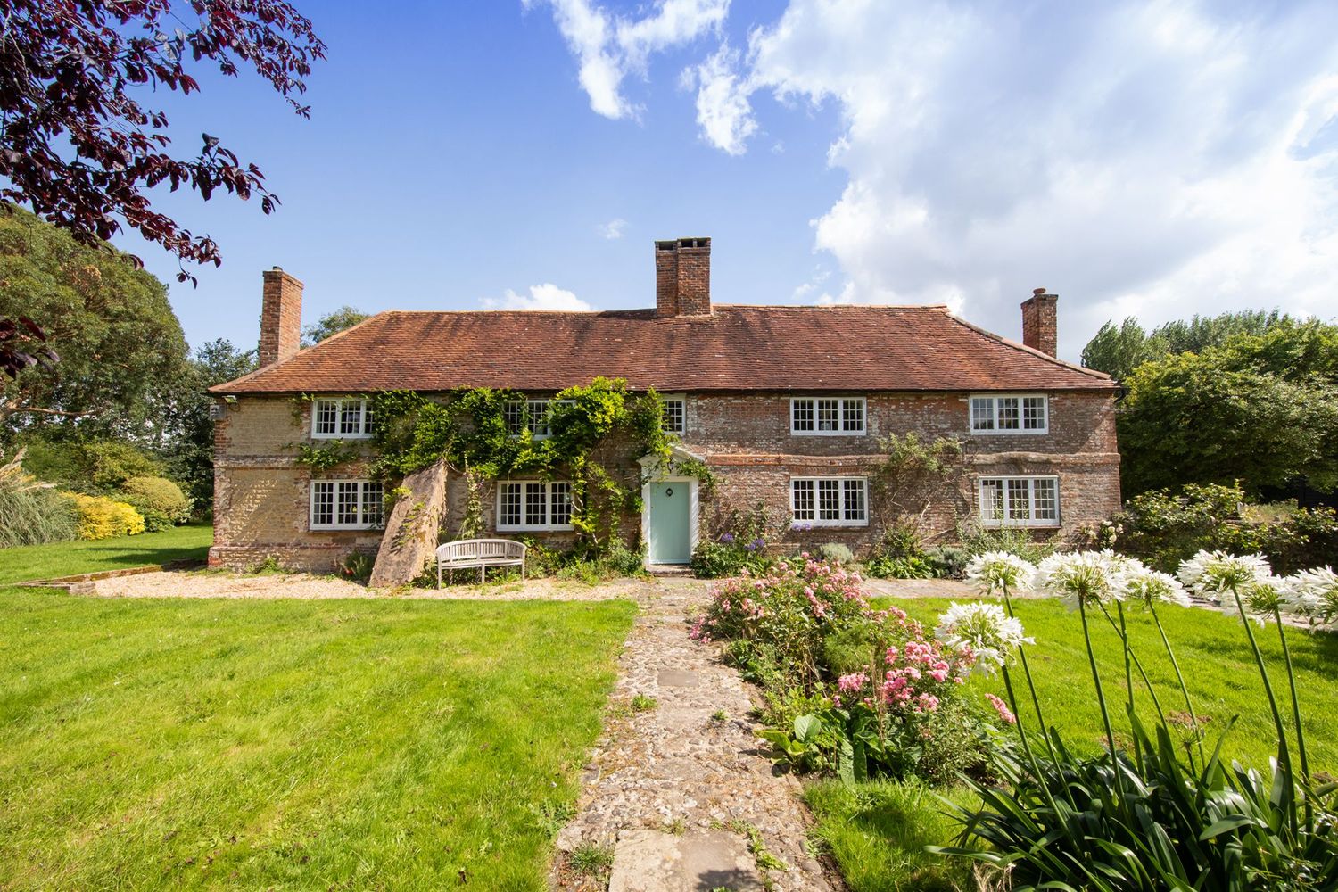 Charming country cottage with red tile roof and lush garden surrounded by mature trees.