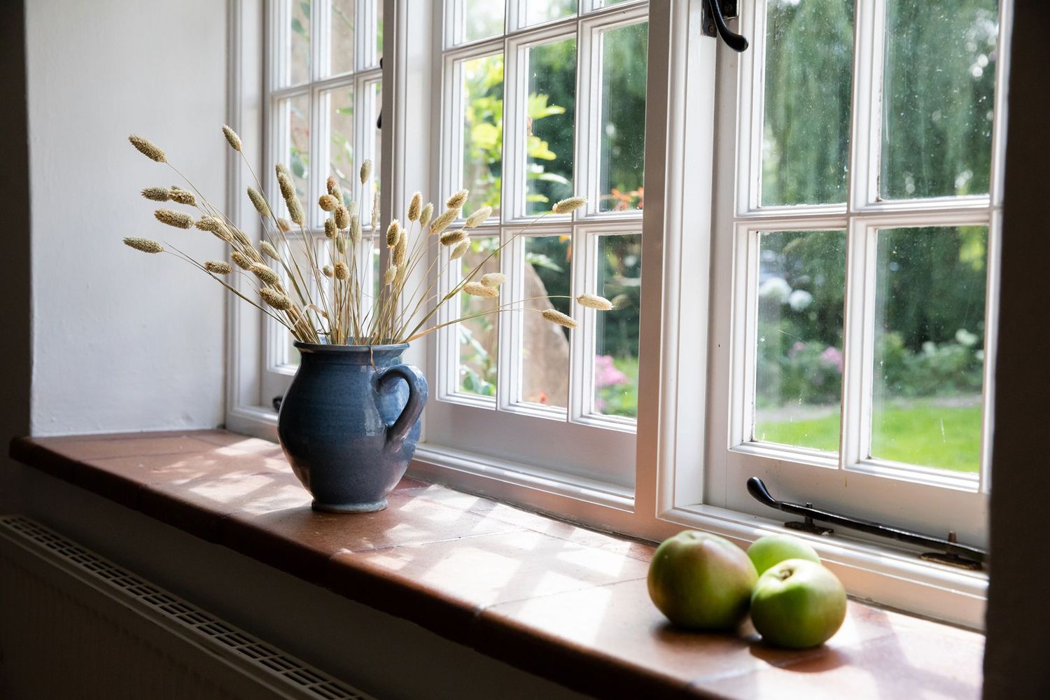 Windowsill decorated with blue ceramic vase of dried flowers and green apples in natural light.