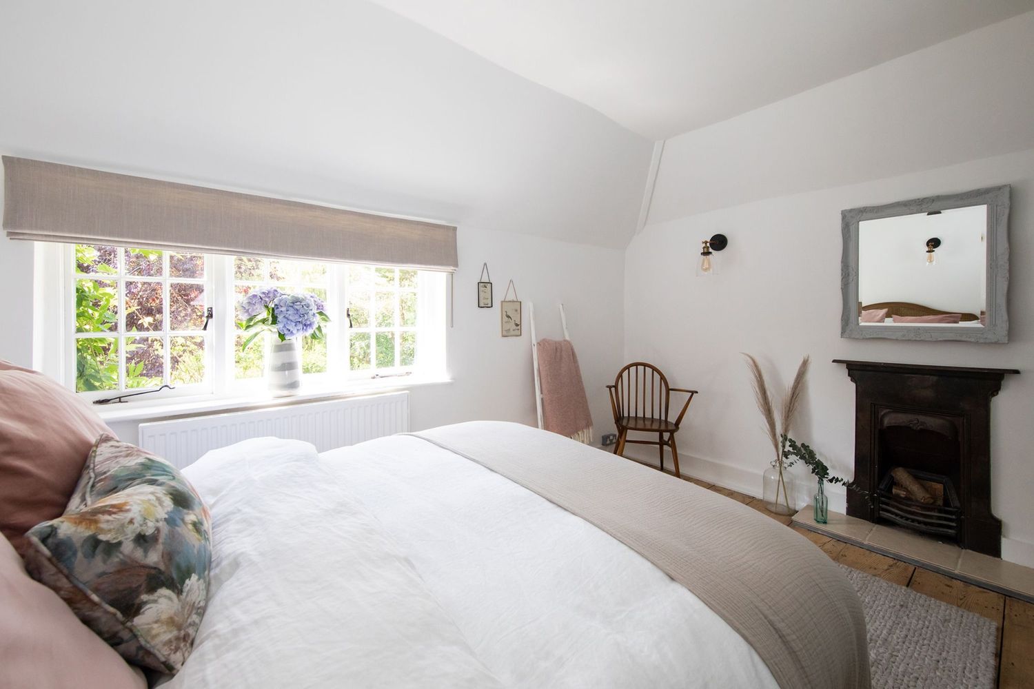 Bright bedroom with large window, white bedding, and traditional fireplace feature.