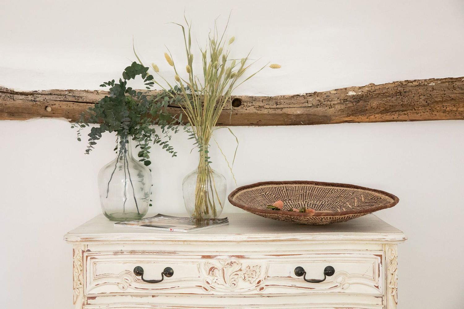 Distressed white dresser topped with glass vases and dried flowers beneath rustic wooden beam.