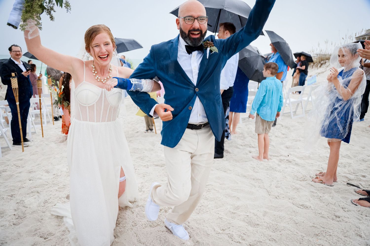Joyful couple celebrates their beach wedding by jumping in the sand while guests look on and applaud.