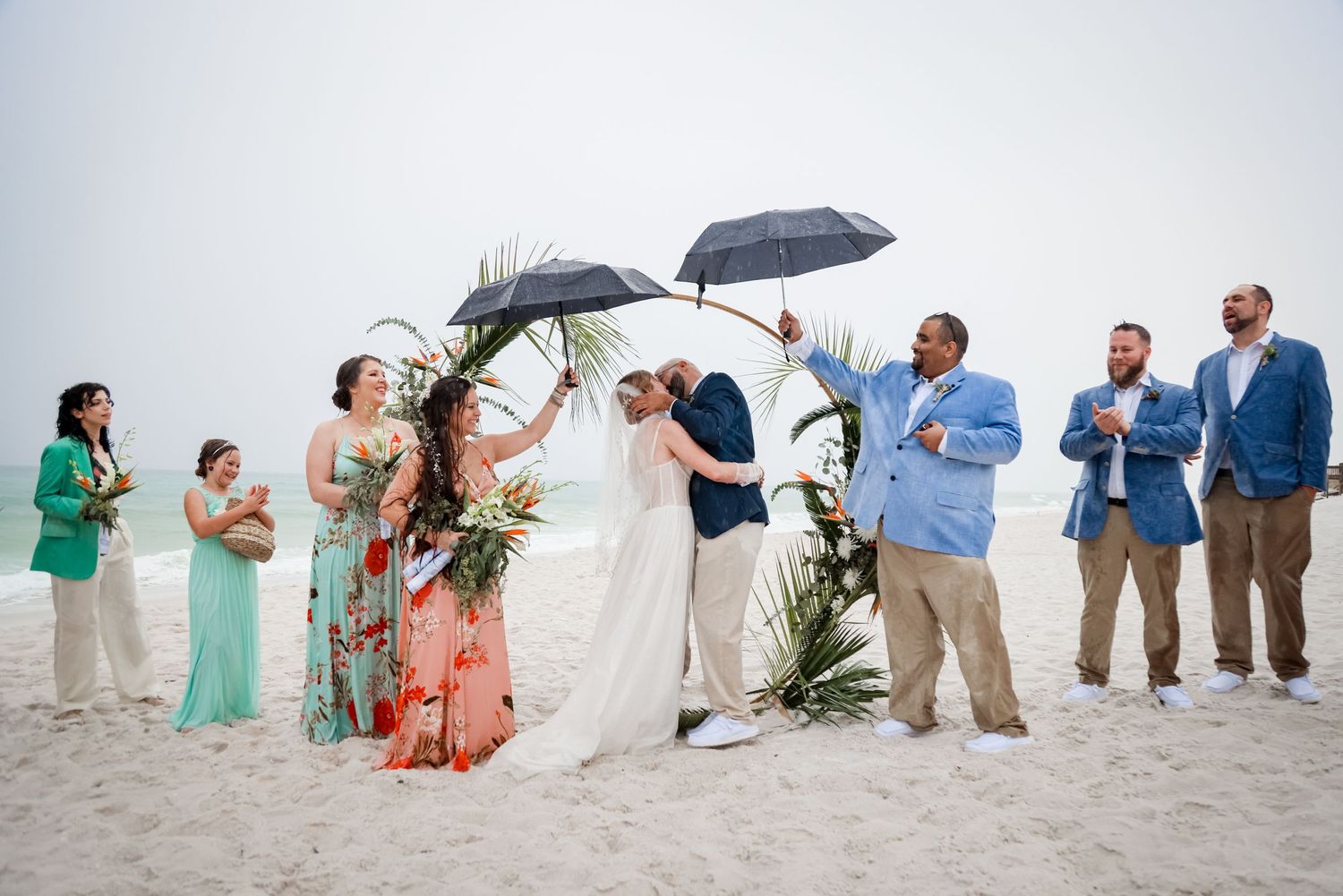Wedding ceremony taking place on a beach with attendees holding umbrellas on an overcast day.