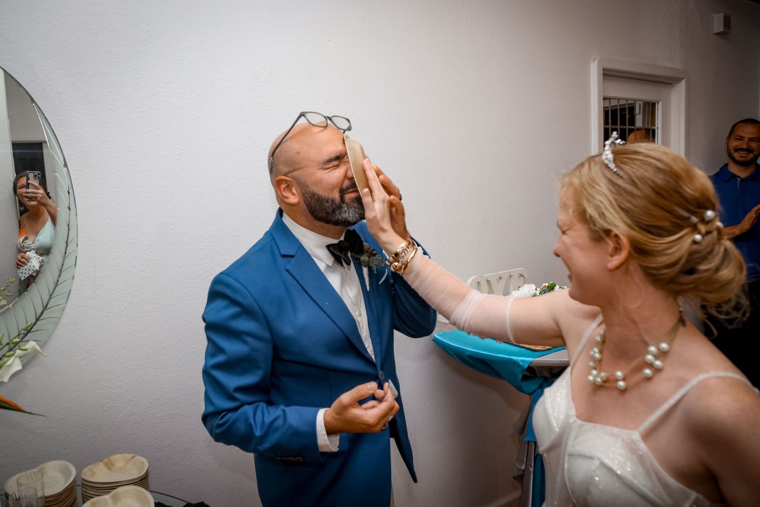 Bride playfully smudges frosting on groom's face during their wedding reception while wearing a blue suit and white dress.