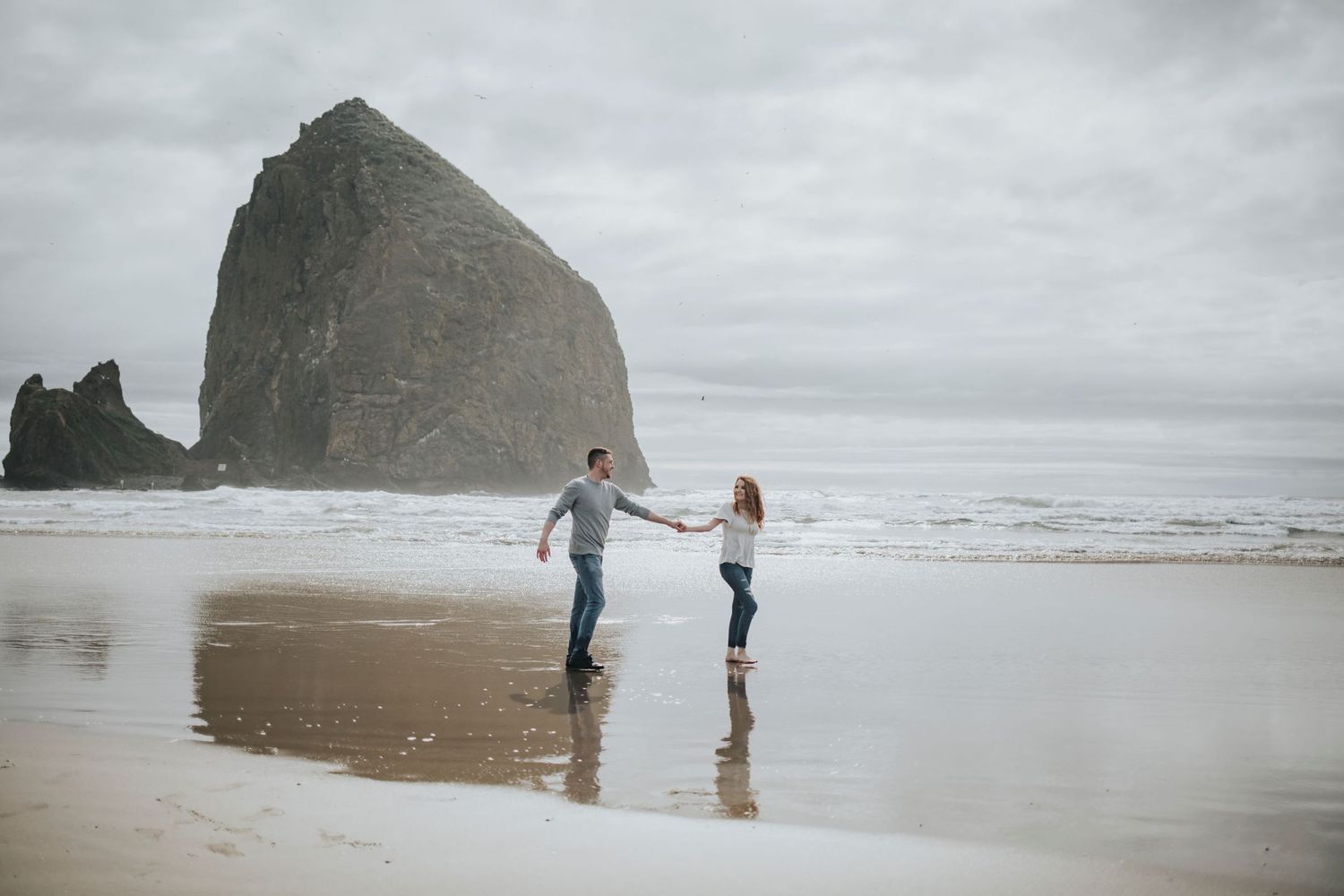 Girl and Boy holding hands walking on the Oregon coast in front of large rock at Cannon beach