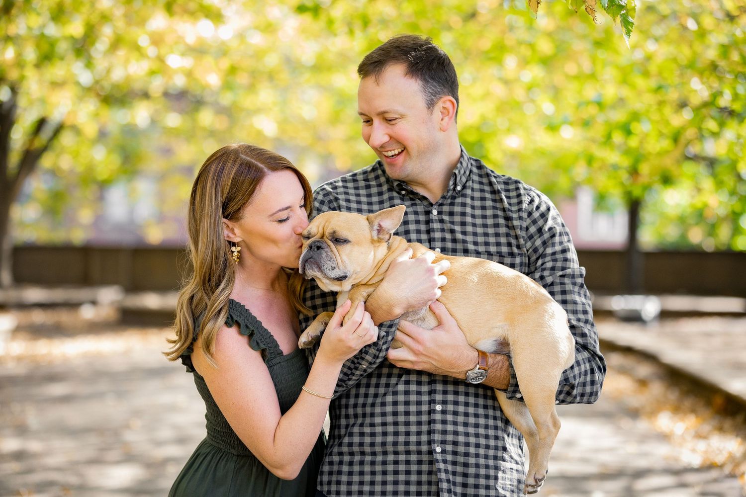 A happy couple holds their small tan dog in a sunny park setting.