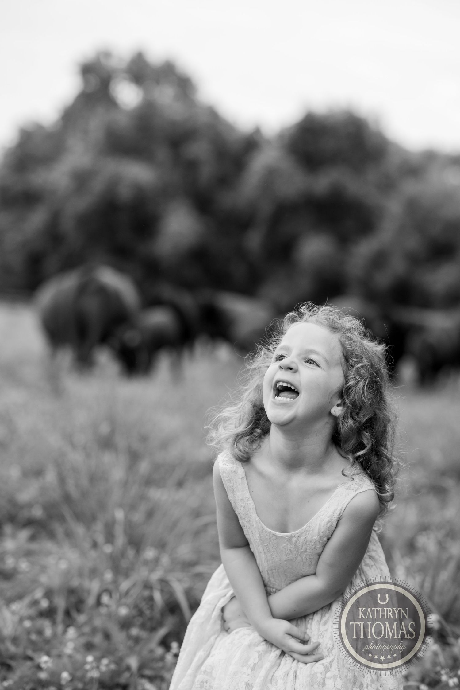 A joyful child in a white dress laughs heartily in a sunny field with cattle in the background.