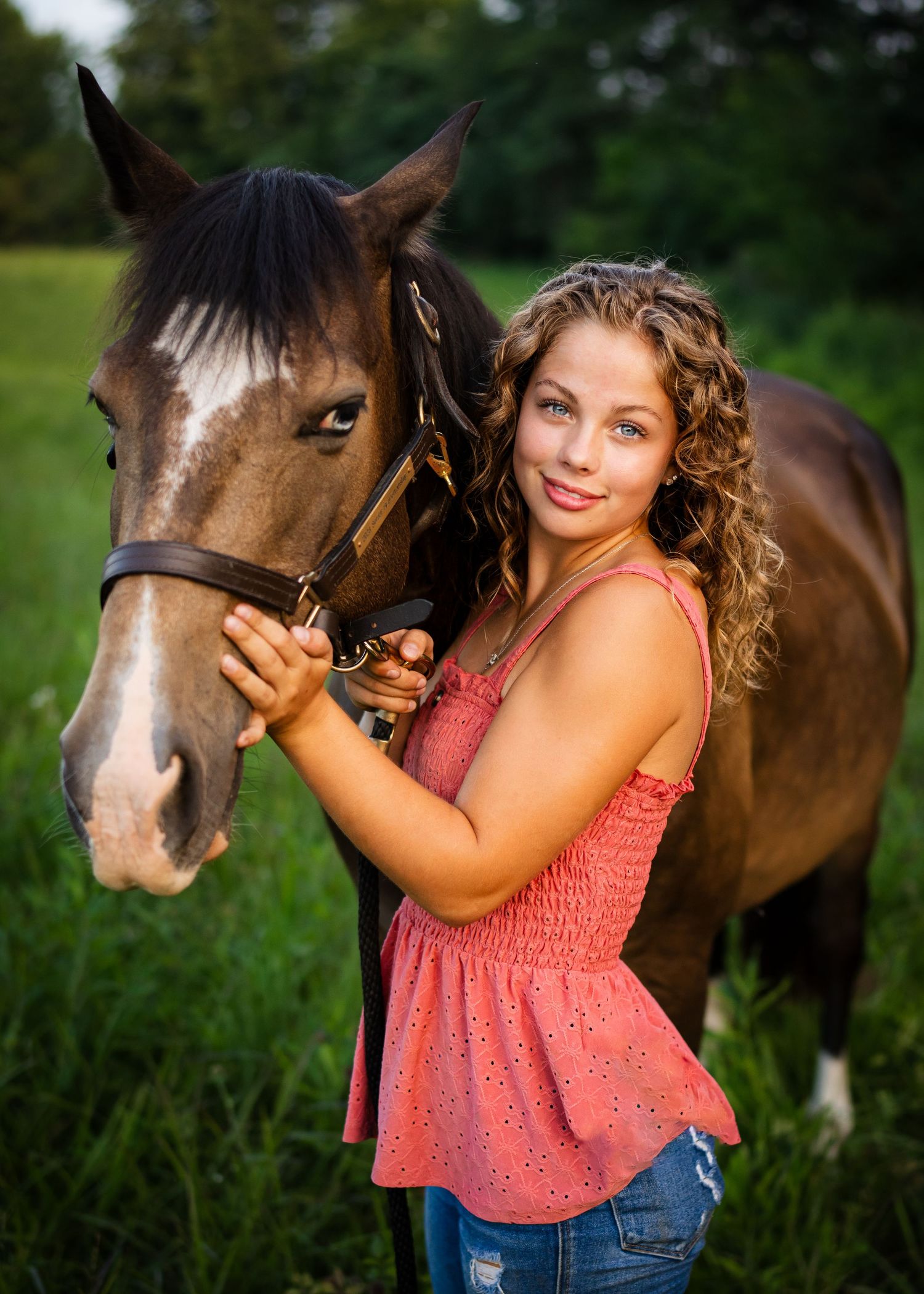 Young woman in coral top poses with brown horse in summer field.