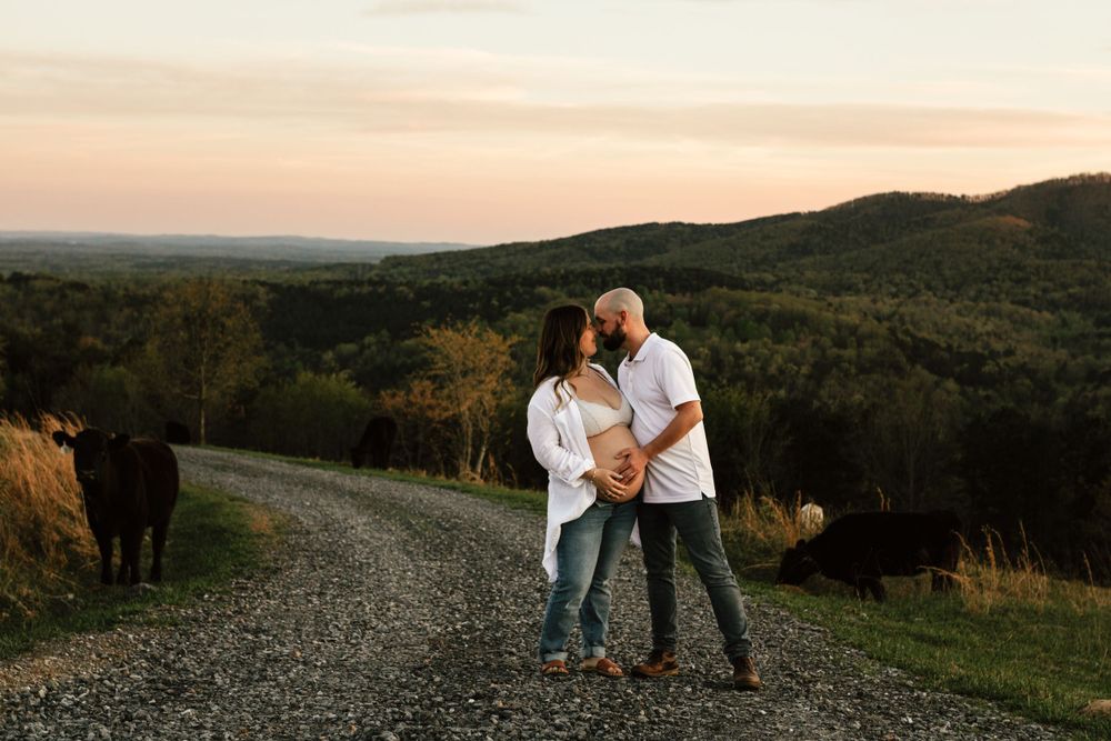 Expecting couple embraces on dirt road with rolling hills at sunset.