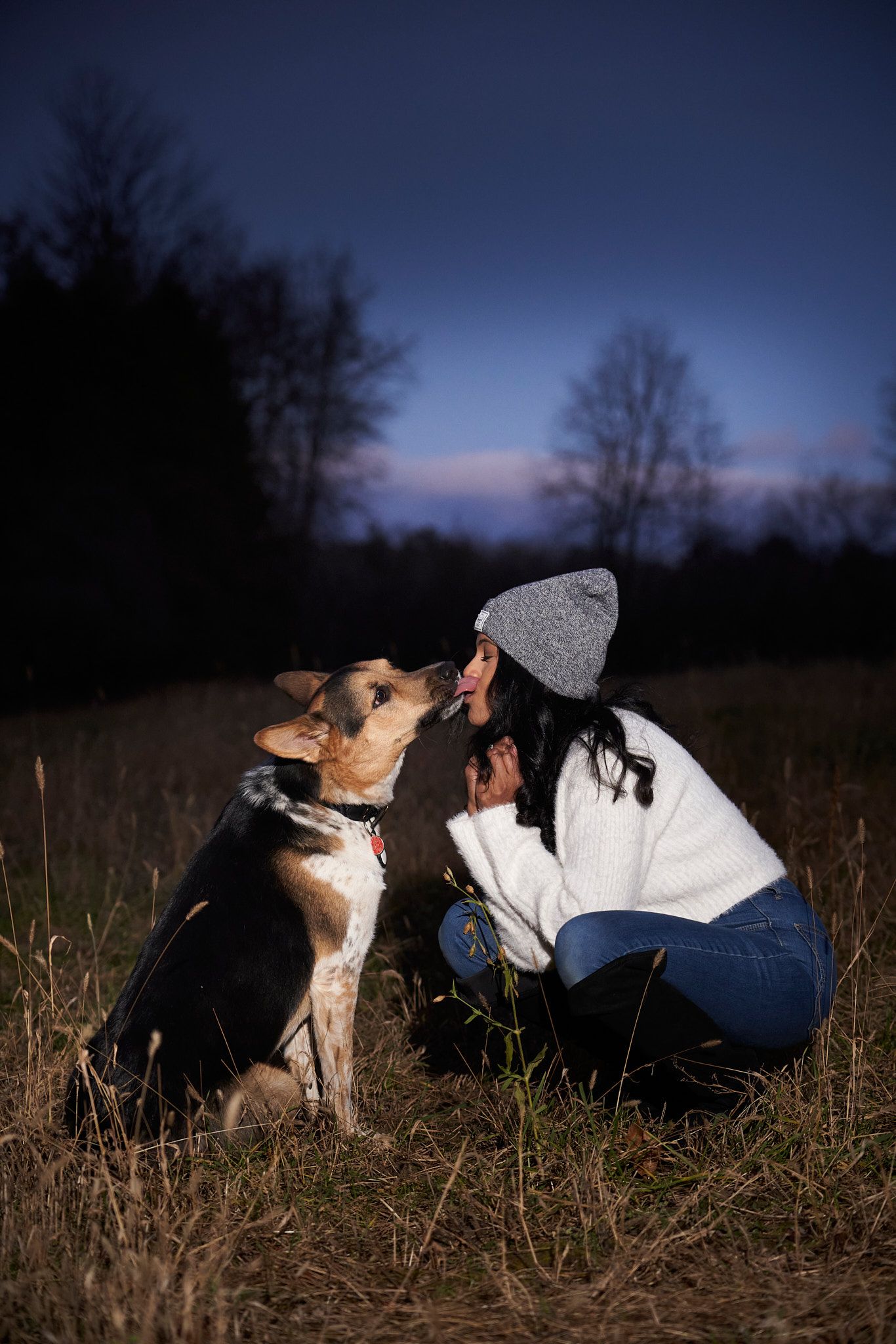 A person in a grey beanie and white sweater crouches in a field at dusk with their mixed breed dog as the dog gives her kisses.
