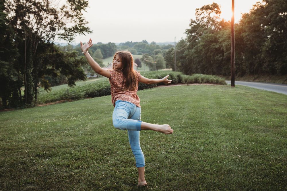 A joyful dance pose against a sunset landscape while wearing jeans and a patterned top.
