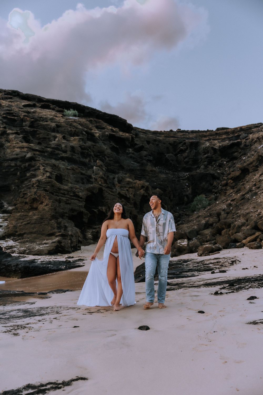 Couple walking along rocky beach shoreline in flowing white dress and casual attire.
