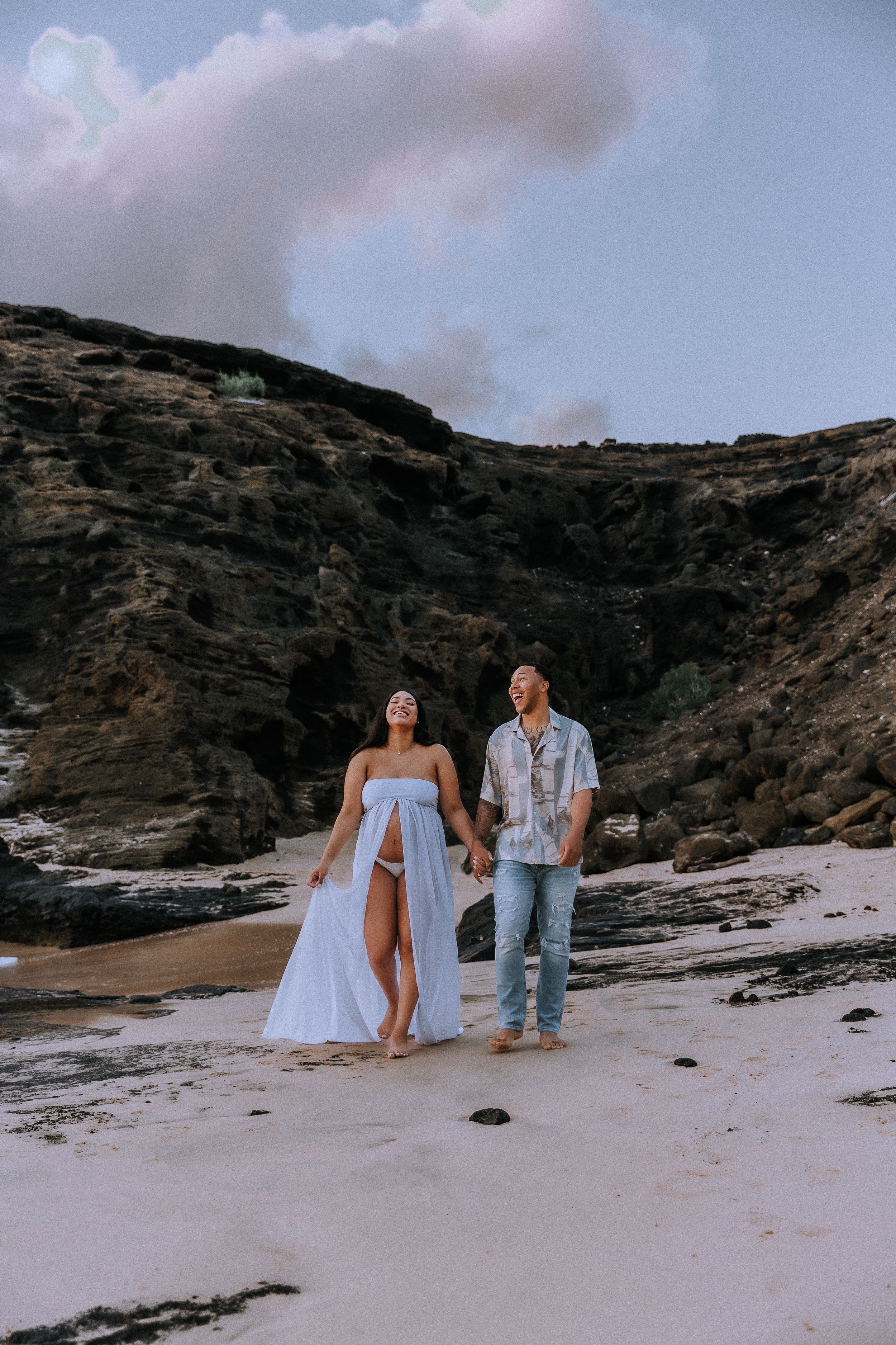 Couple walking along rocky beach shoreline in flowing white dress and casual attire.
