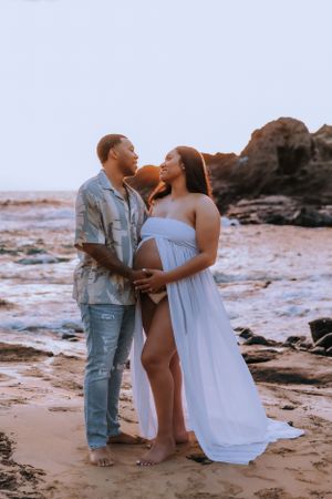 A maternity photoshoot on a beach at golden hour with rocky cliffs in background.