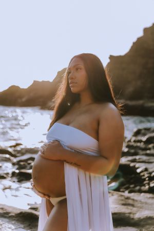 Maternity photoshoot at sunset on a rocky beach with flowing white fabric and gentle lighting.