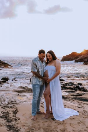 A romantic maternity photoshoot on a rocky beach at sunset with a couple in casual attire.