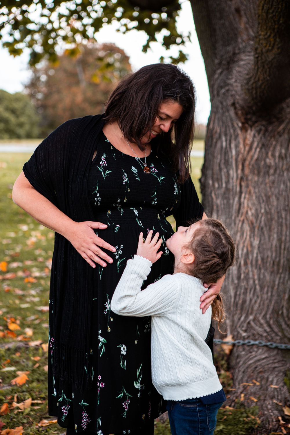 Child reaches up to touch mother's pregnant belly under autumn tree.