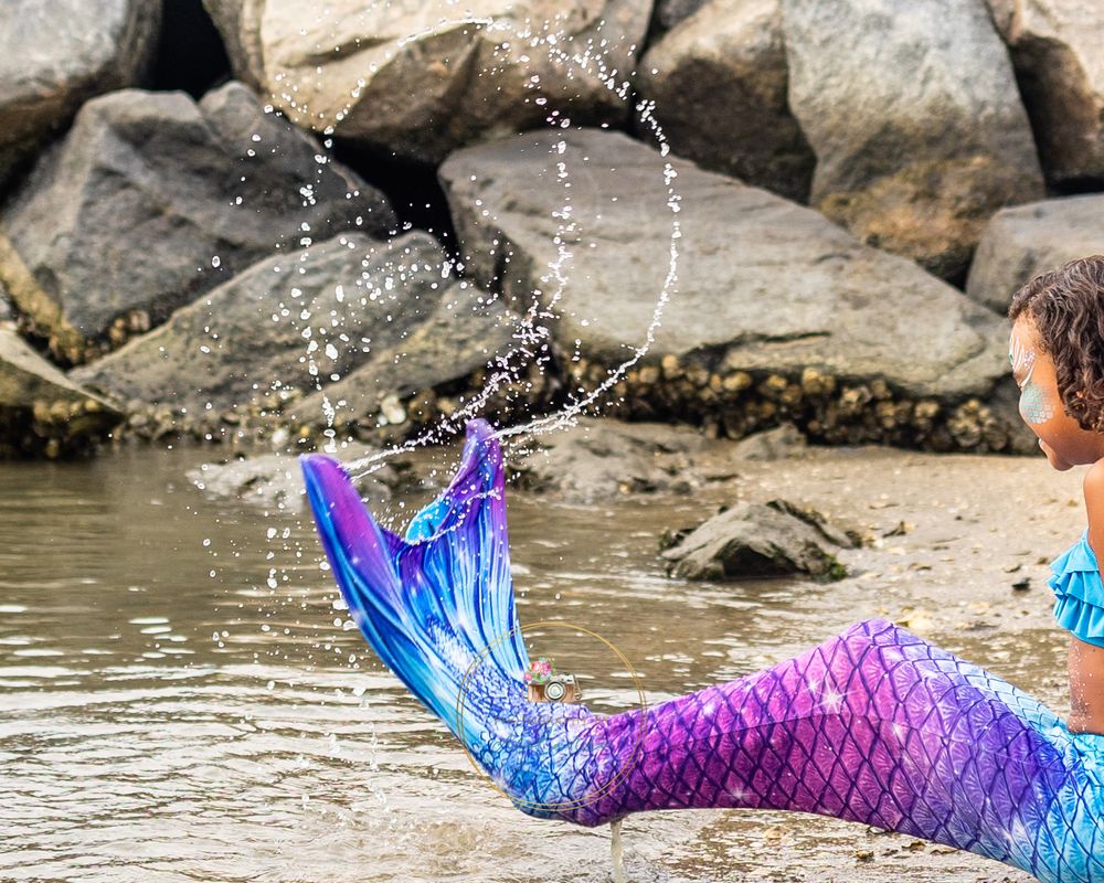 Mermaid tail splashing water against rocky shoreline in playful photo.