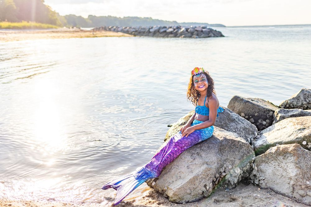 A costumed mermaid in purple and blue tail sits on rocky shoreline during golden sunset.