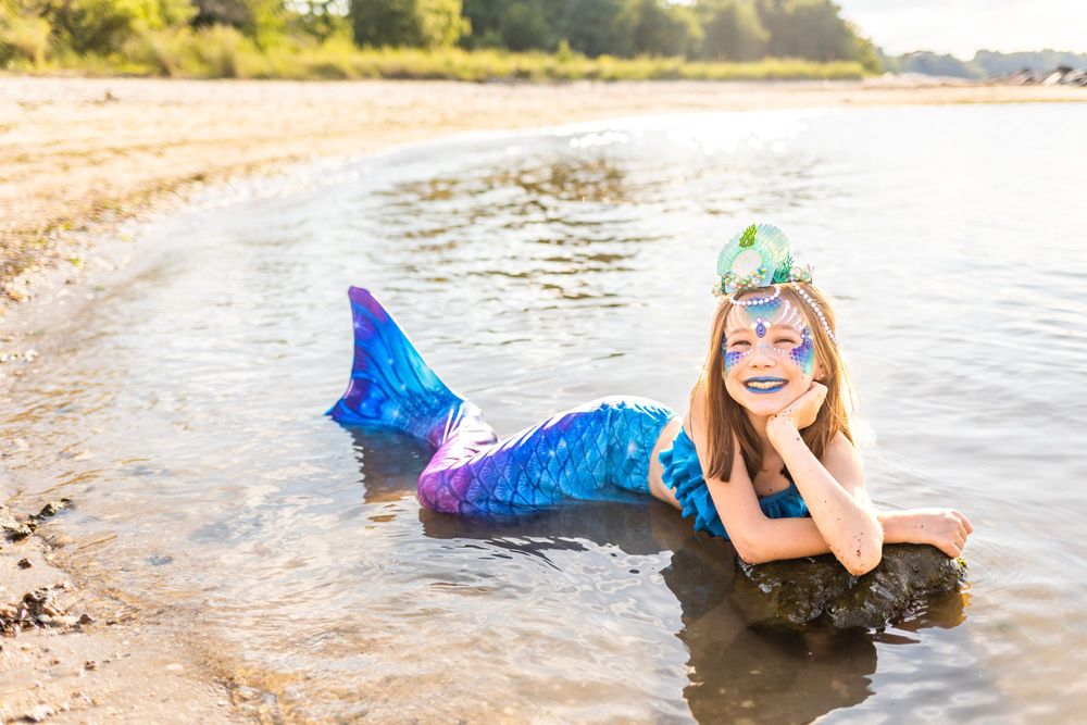 A mermaid-costumed figure poses on a beach shoreline in a blue tail costume with face paint and green crown.