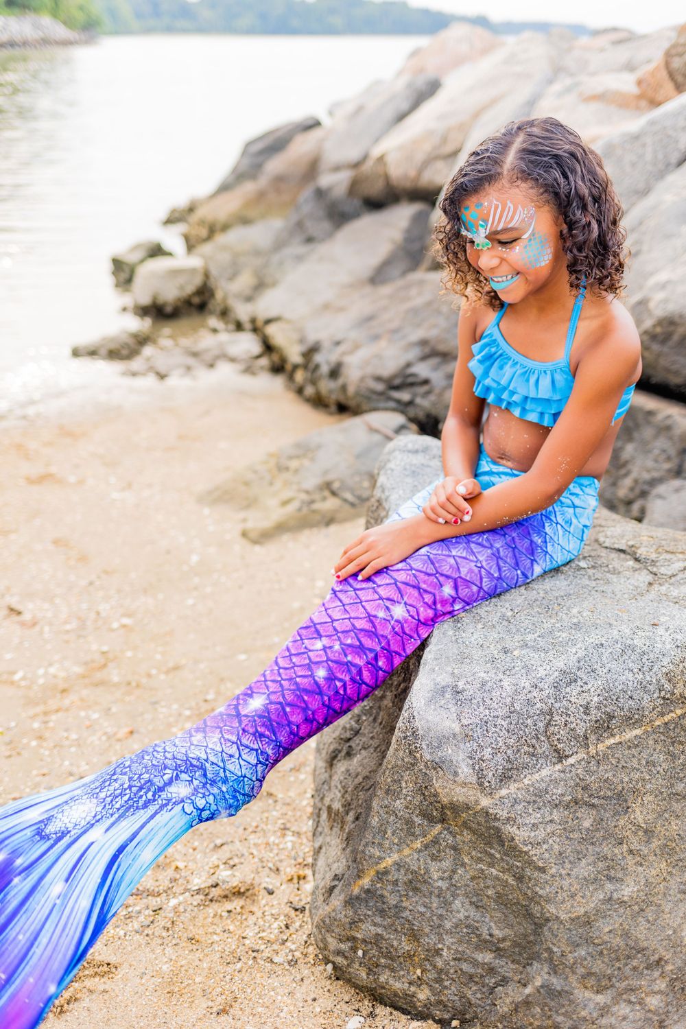 Mermaid costume wearer with decorated face perched on coastal rocks by water.