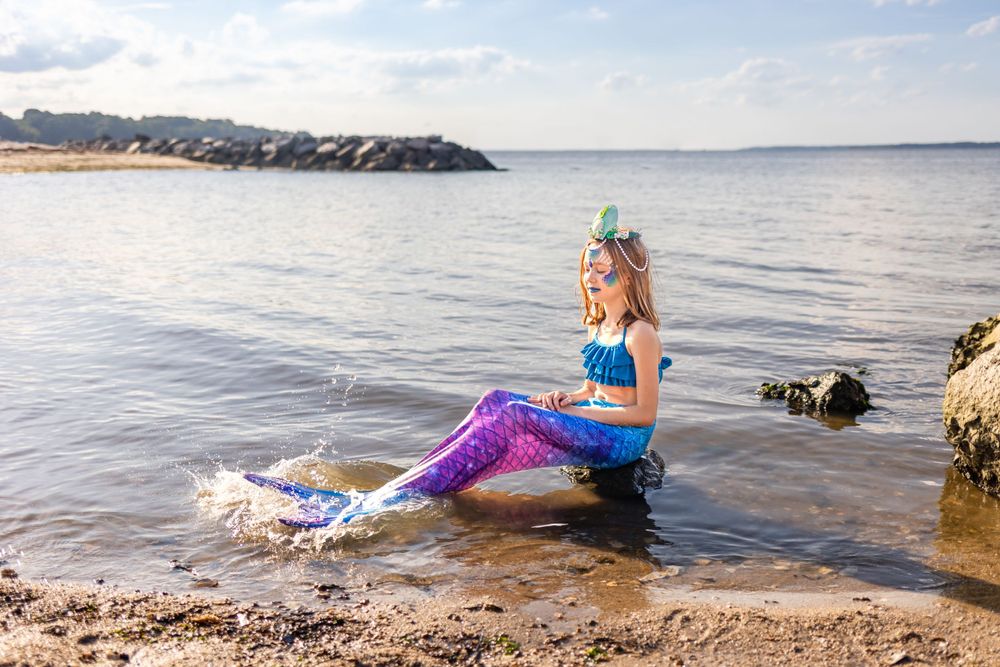 Costumed performer in purple mermaid tail splashing in shallow coastal waters.