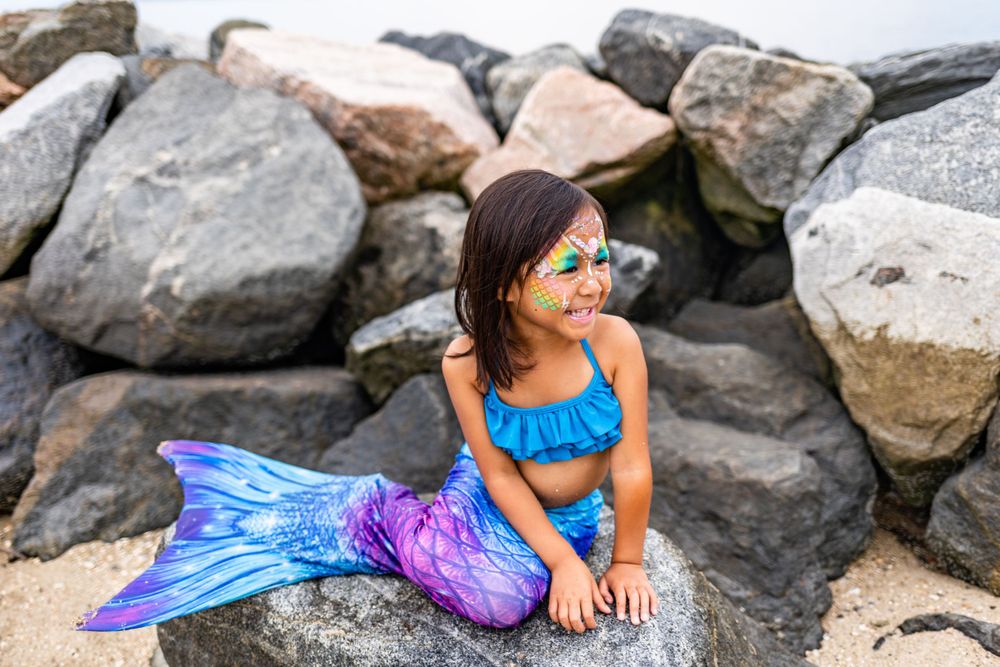 Performer in bright mermaid costume posing on rocky beach with ocean backdrop.