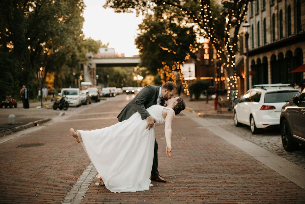 Groom dipping bride in white gown on brick street at sunset with string lights.