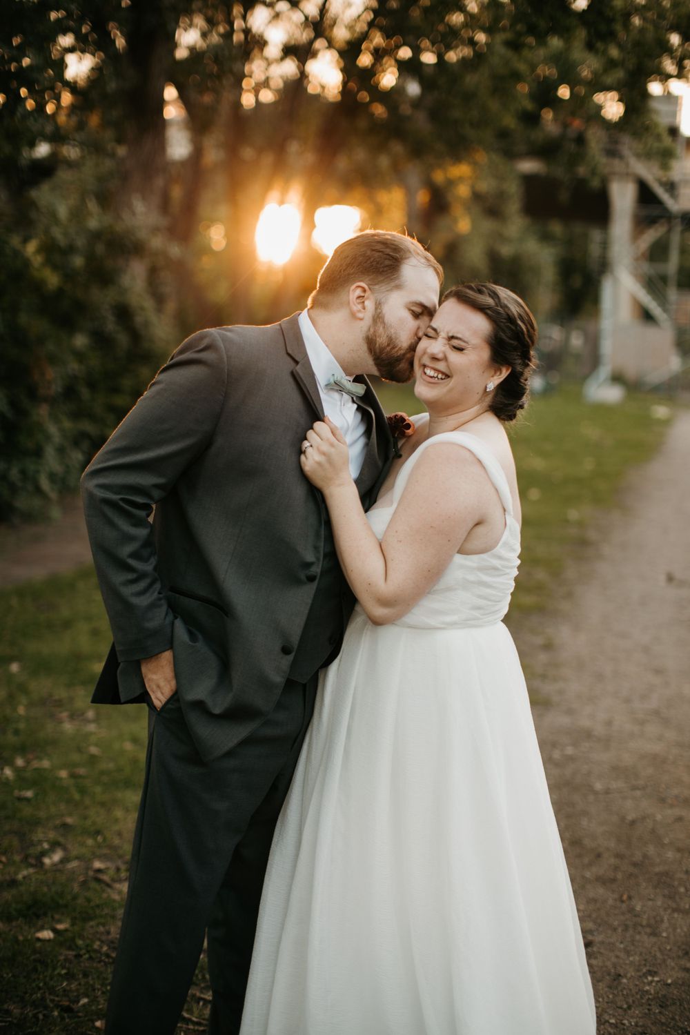 Groom in gray suit kissing bride on cheek during golden hour outdoor wedding portrait.