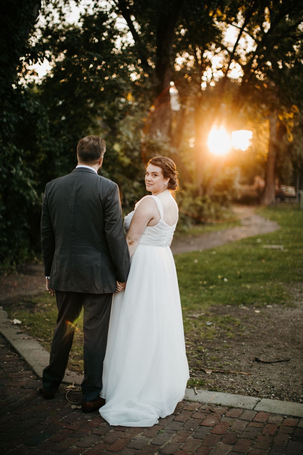 Bride in white gown glances back while holding grooms hand on brick path at sunset.