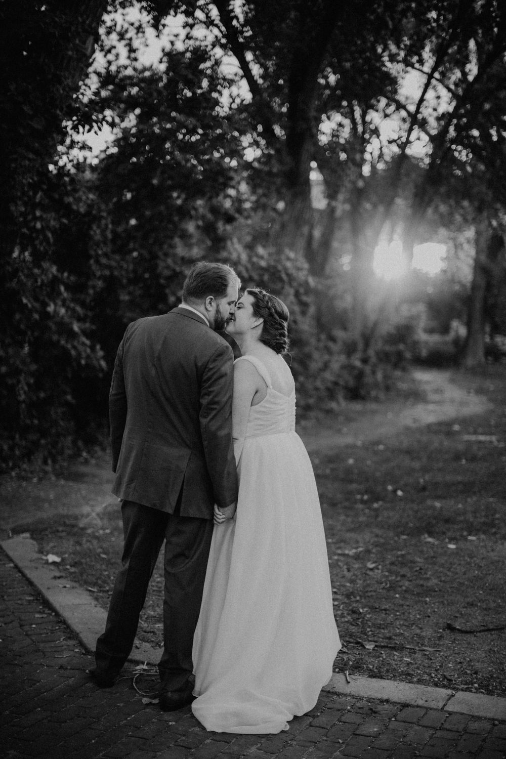 Black and white photo of bride and groom kissing on brick path with sunlight behind.