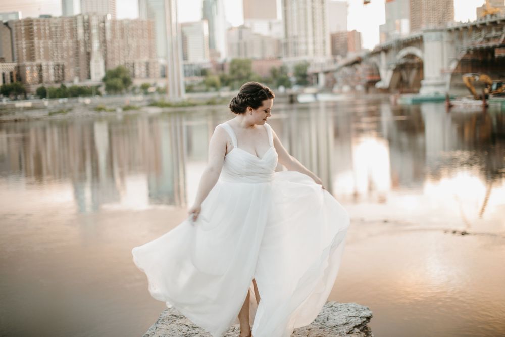 Bride twirling white wedding gown on rocks by river with city skyline at golden hour.